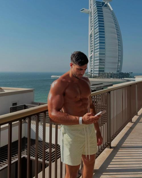 Shirtless man using phone on balcony overlooking ocean and Burj Al Arab.