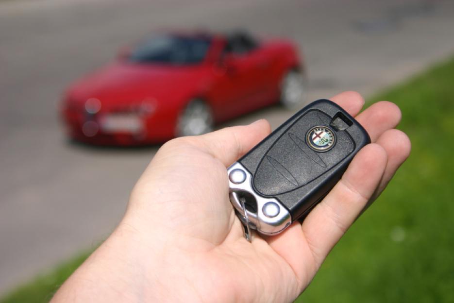 Hand holding an Alfa Romeo car key with a red Alfa Romeo Spider in the background.