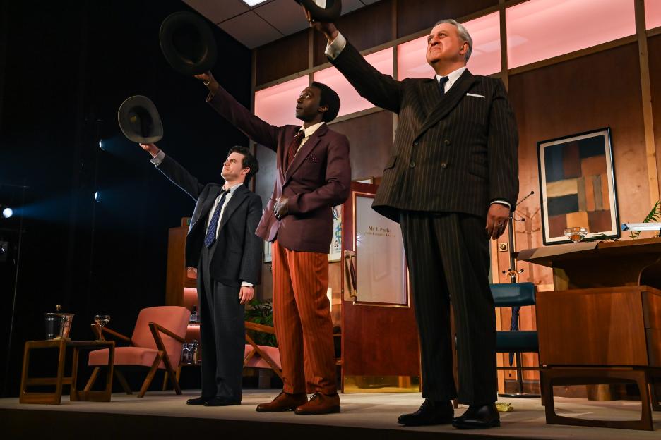 Three actors in 1950s suits on a stage, raising their hats.