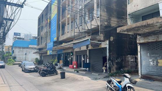 Street scene in Bangkok, Thailand, with parked cars and motorcycles near shops and buildings.