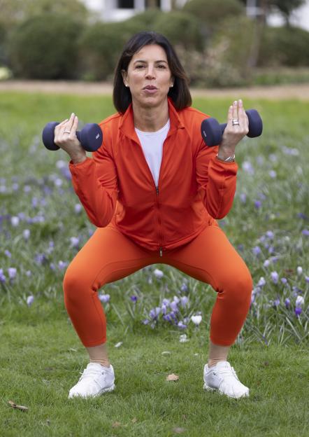 Woman in orange workout clothes squatting while lifting weights outdoors.