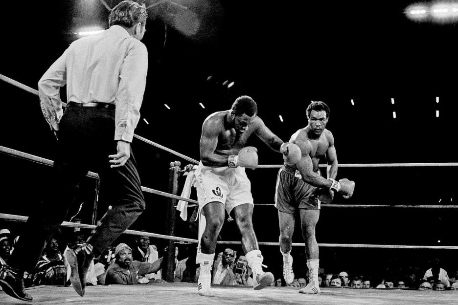 Black and white photo of Joe Frazier being hit by George Foreman in a boxing match.