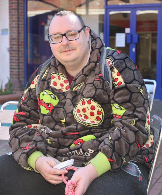 A man wearing a Teenage Mutant Ninja Turtles and pizza-patterned fleece sits outdoors holding a dog leash and a cup.