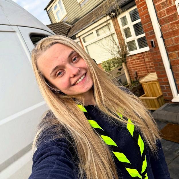 Young woman with long blonde hair, wearing a dark blue fleece and a neon yellow and black neckerchief, standing in front of a van and house.