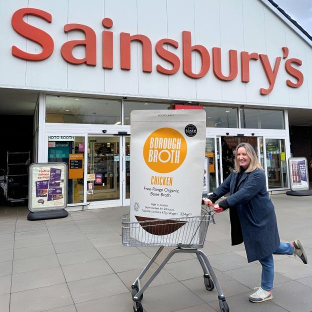 Woman pushing shopping cart with Borough Broth packaging in front of Sainsbury's.