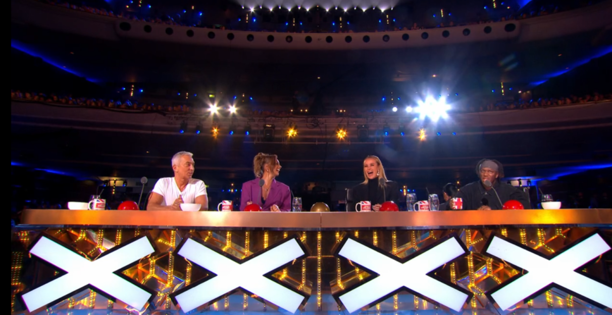 Screenshot of four judges sitting at a judging table with large X's on the front.