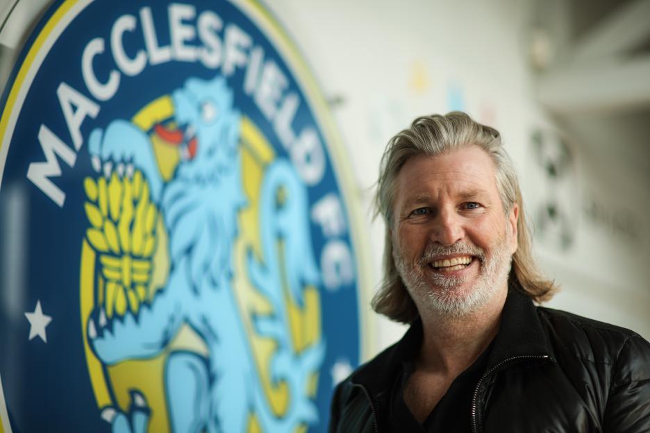 Robbie Savage, Director of Football at Macclesfield FC, smiling in front of the team's logo.