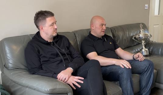 Two men sitting on a couch, one holding a trophy.
