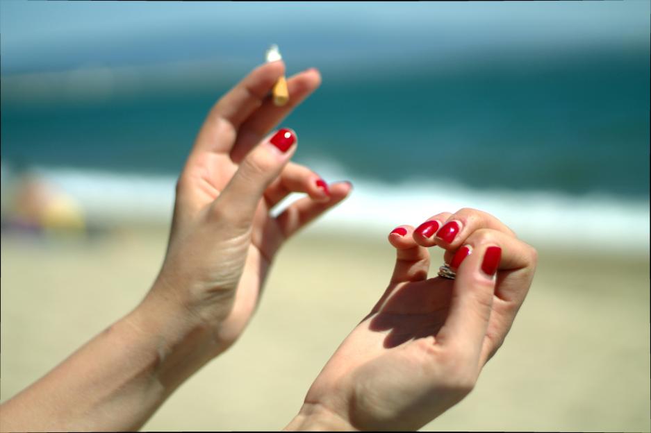 Woman's hands with red nail polish holding a cigarette on a beach.