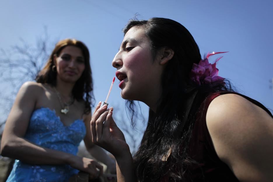 A young Romani woman applying lip gloss, with another woman in the background.