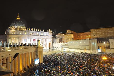 Nighttime view of St. Peter's Square at the Vatican, filled with a large crowd under umbrellas.