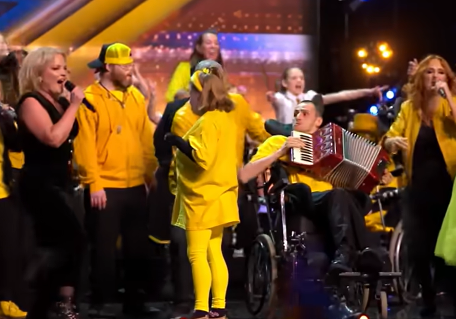 Group of people in yellow singing and playing music, one person in a wheelchair plays an accordion.