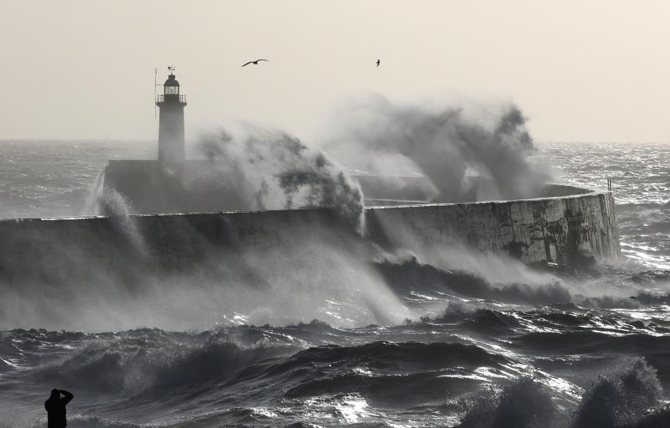 Person observing large waves crashing against a sea wall and lighthouse during a storm.