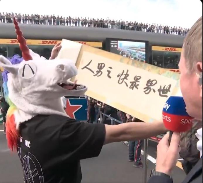 Person in a unicorn mask holds a sign with Chinese characters and speaks to a reporter.