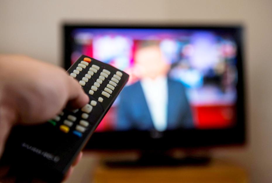 Hand holding a TV remote with a blurred television screen in the background.