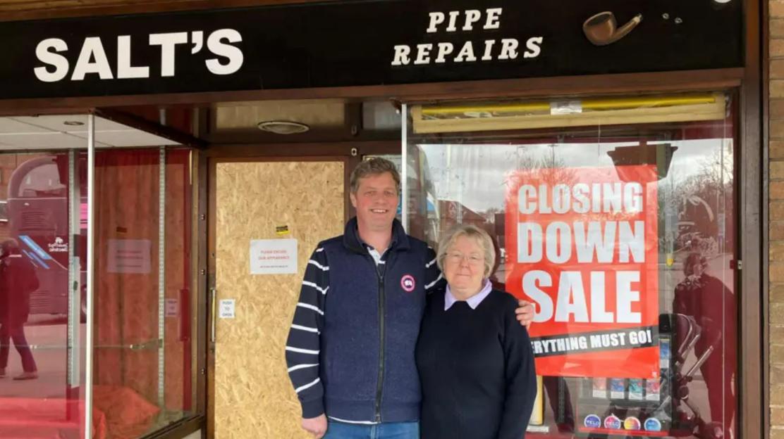 A man and woman stand in front of Salt's Pipe Repairs, which is having a closing down sale.