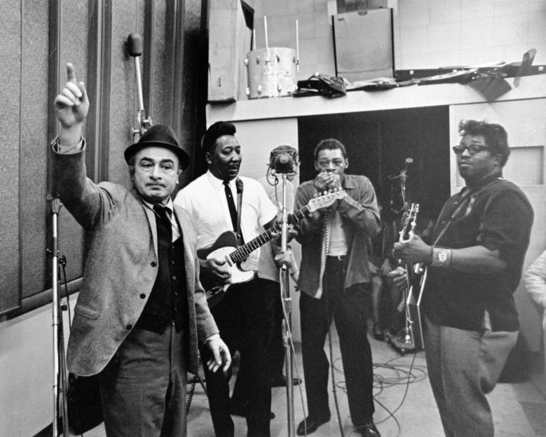 Black and white photo of Phil Chess supervising Muddy Waters, Little Walter, and Bo Diddley recording in a studio.