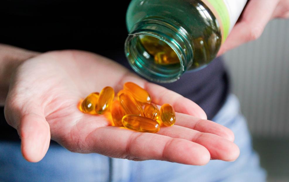 Hand holding several yellow vitamin D and flaxseed oil capsules poured from a bottle.