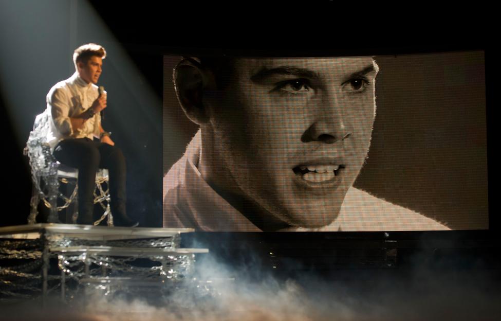 Aiden Grimshaw on stage during The X Factor, with a large screen behind him showing a close-up of his face.