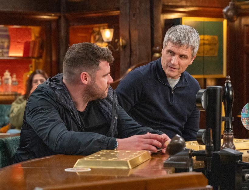 Two men talking at a bar, one looking concerned.