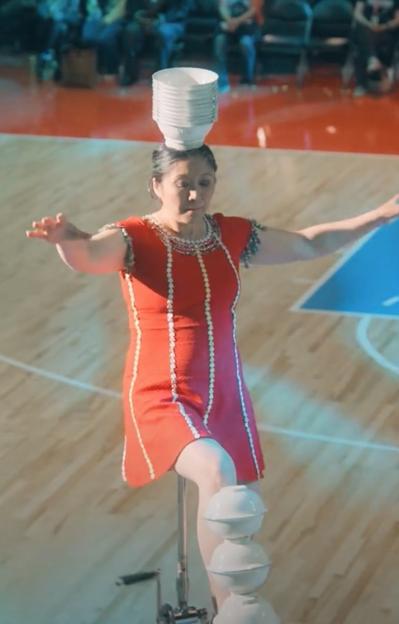 Woman in red dress balancing bowls on her head and legs while unicycling.