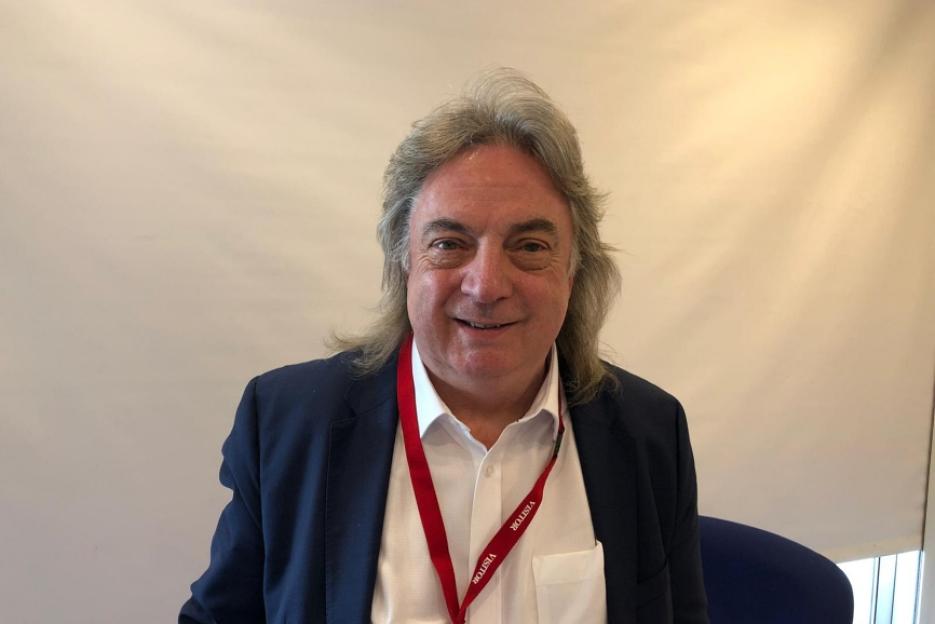 Portrait of a man with long gray hair wearing a navy blue blazer and a red lanyard.