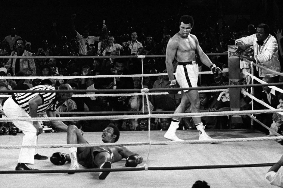 Black and white photo of a boxing match; a boxer is down on the canvas while the other boxer stands over him.