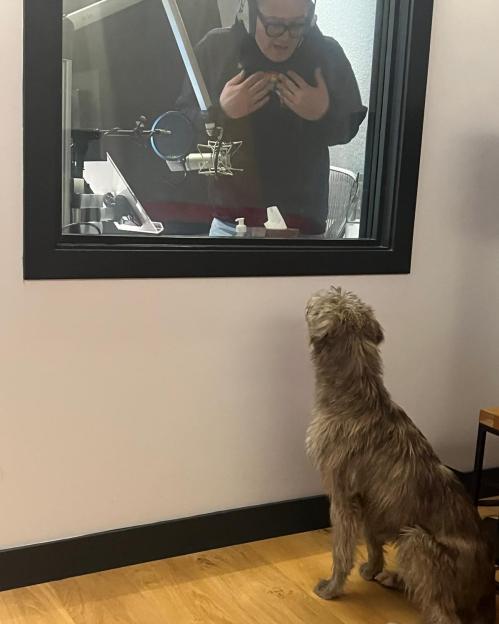 Alan Carr in a recording studio, viewed through a window, with a dog sitting outside.