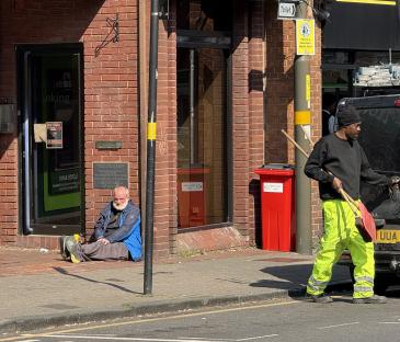 A street scene showing a man sitting on the sidewalk and a street cleaner walking by.