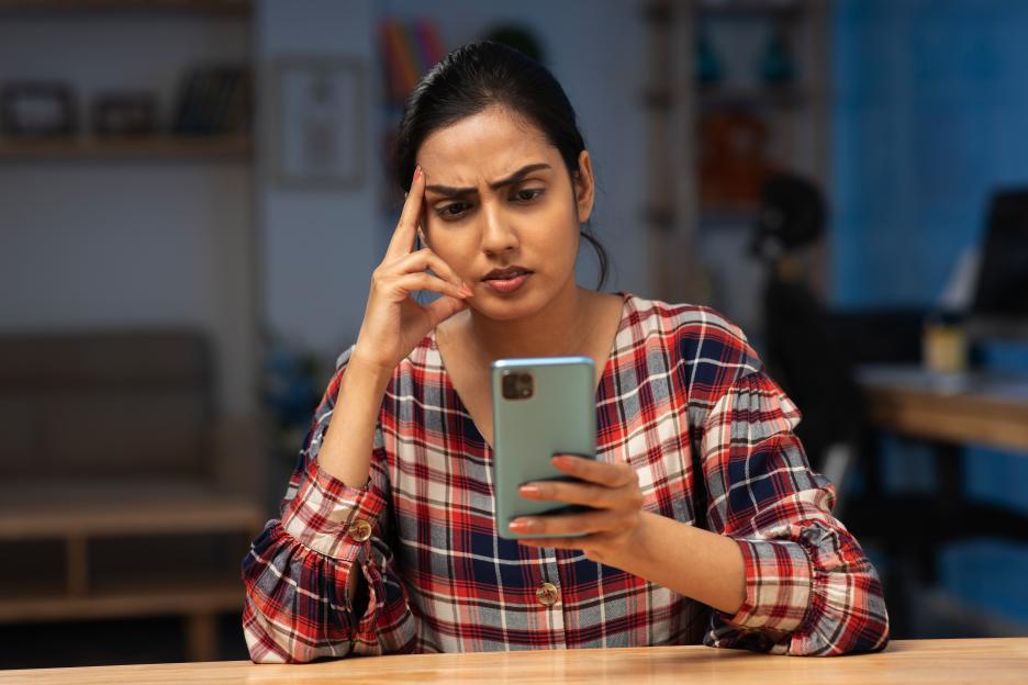 A young Indian woman sits at a table, looking worried as she reads her smartphone.