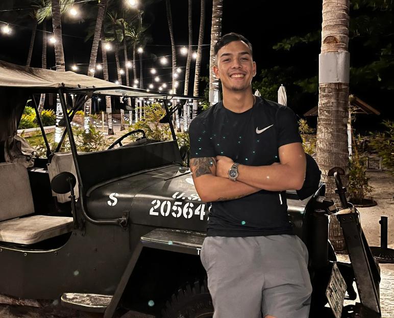 Man standing by a jeep, arms crossed, at night under palm trees with string lights.