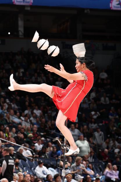 A woman in a red dress unicycling while juggling bowls during a halftime show.