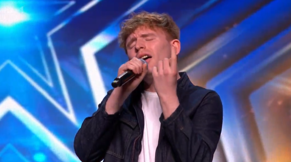 Screenshot of a young man singing into a microphone on a stage with a blue background.