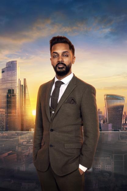 A man in a suit stands in front of a London cityscape at sunset.