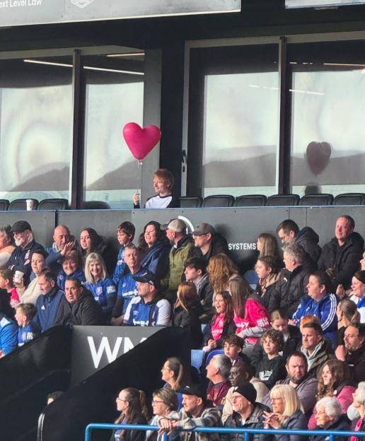 A person holding a heart-shaped balloon stands in front of a crowd of spectators at a stadium.