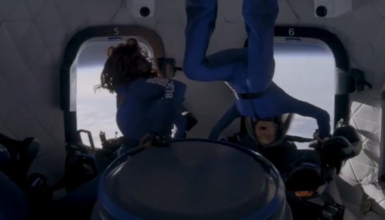 Two women in blue flight suits float upside down in a spacecraft cabin.
