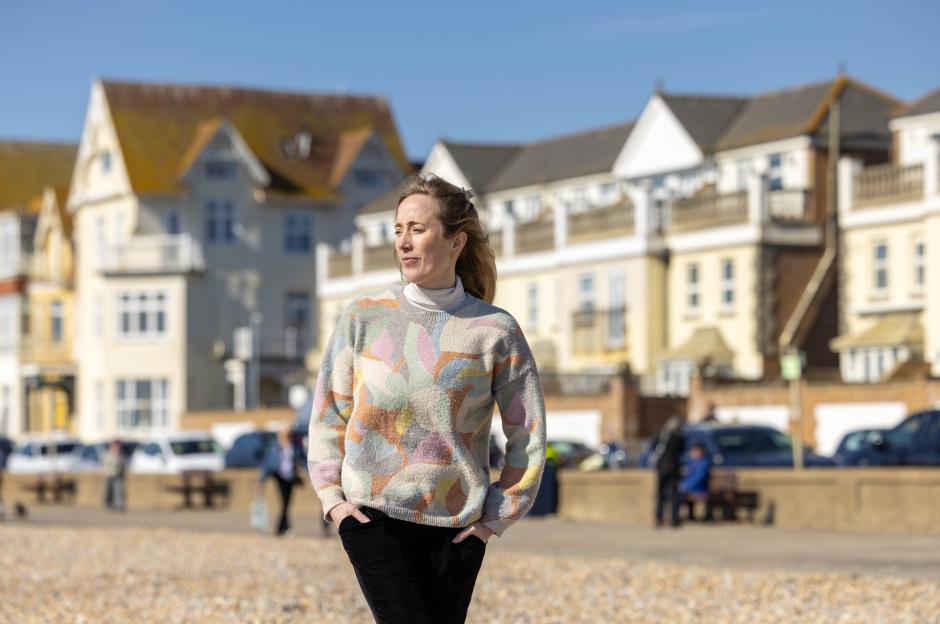 A woman in a patterned sweater stands on a beach with houses in the background.