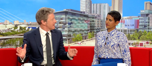 Two news presenters sitting on a red couch, discussing a topic.