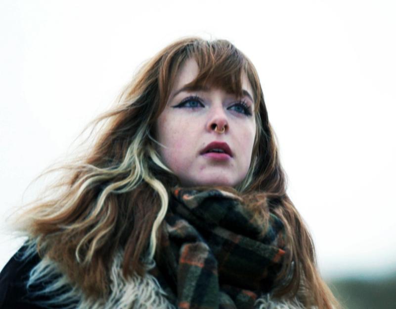 A young woman with long, reddish-brown hair wearing a scarf looks off to the side.