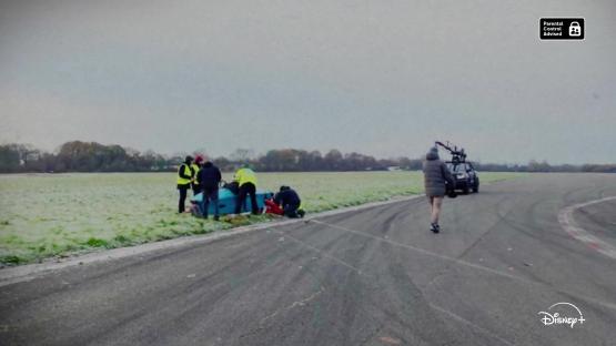 Film crew assisting a person near a flipped vehicle.